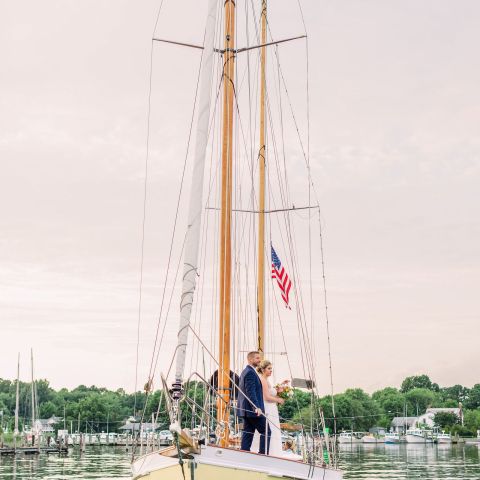 Bride and groom stand on a sailboat with an American flag, calm water and trees in the background.
