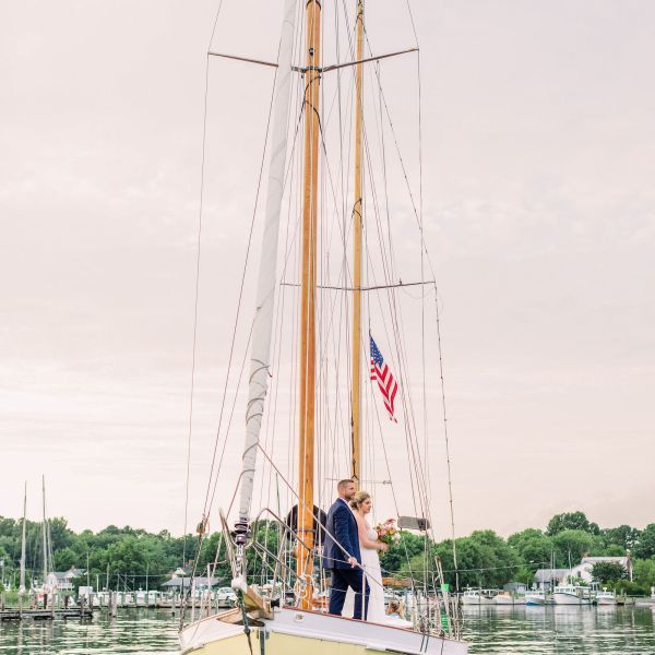 Bride and groom stand on a sailboat with an American flag, calm water and trees in the background.