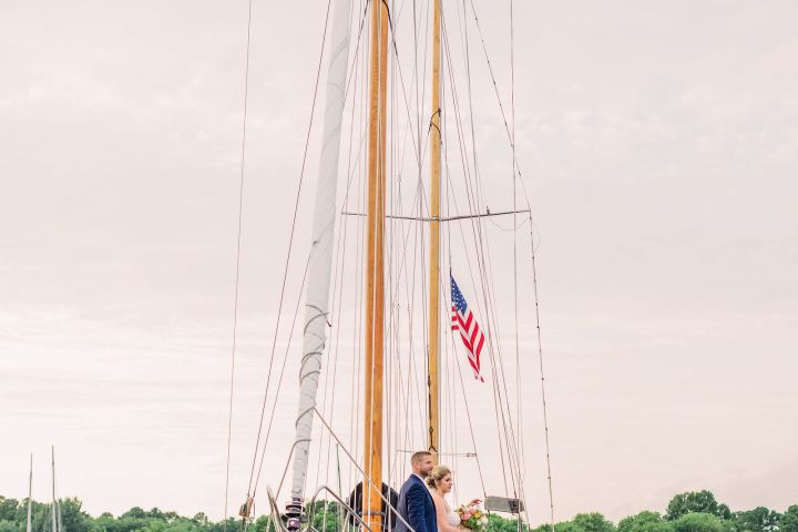 Bride and groom stand on a sailboat with an American flag, calm water and trees in the background.