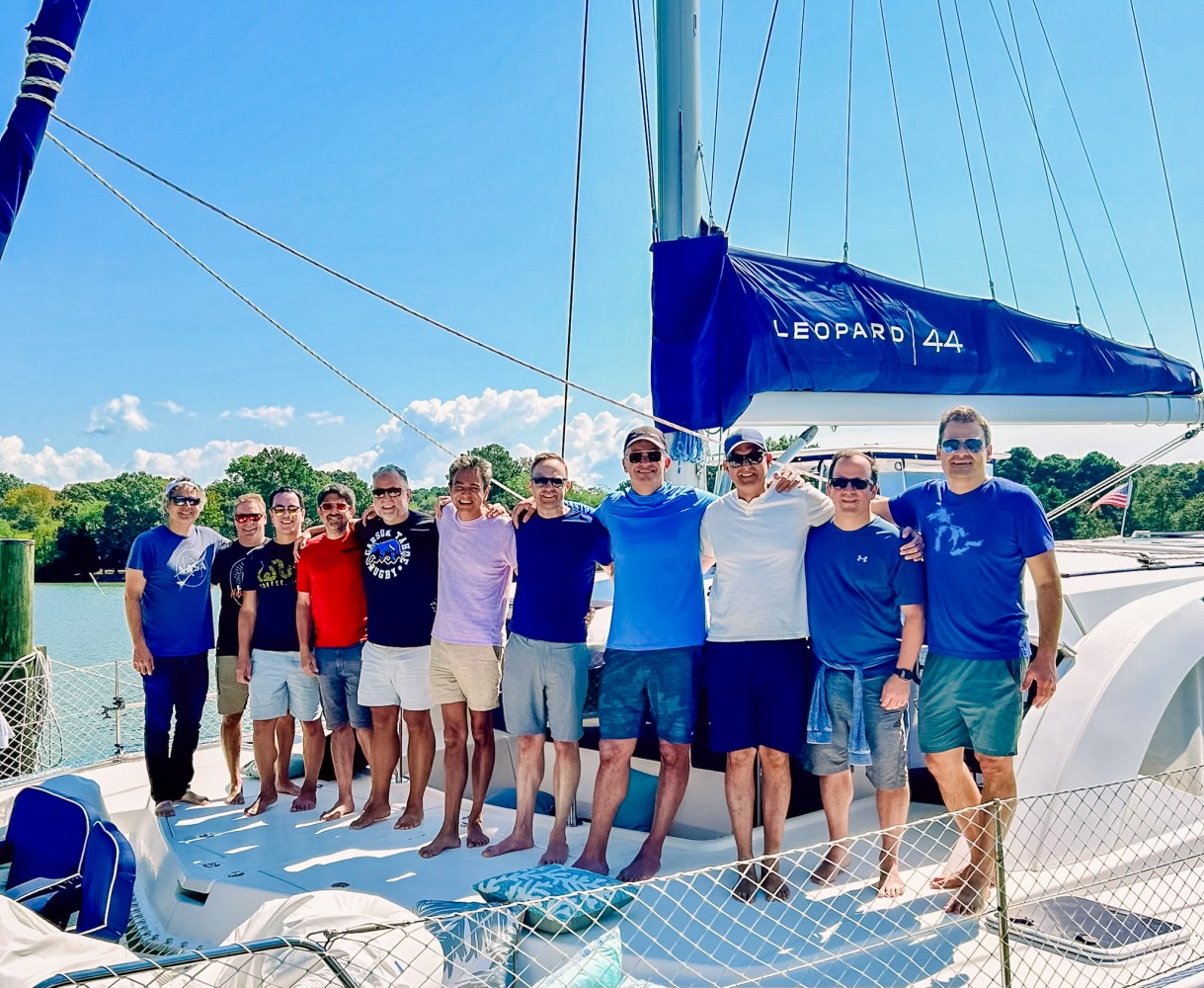 Group of 10 men posing on a Leopard 44 catamaran in a sunny outdoor setting.