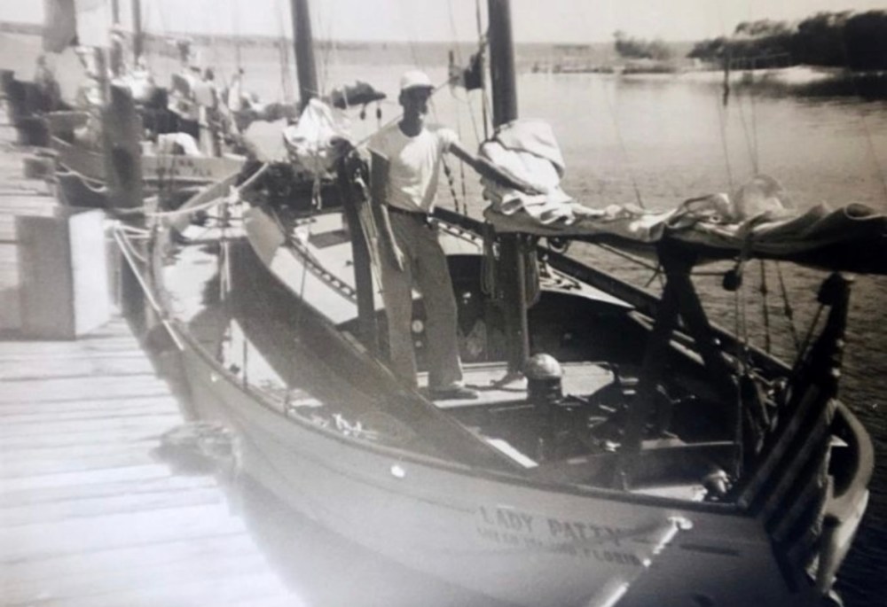 Black and white photo of a person standing on a small sailboat at a dock.