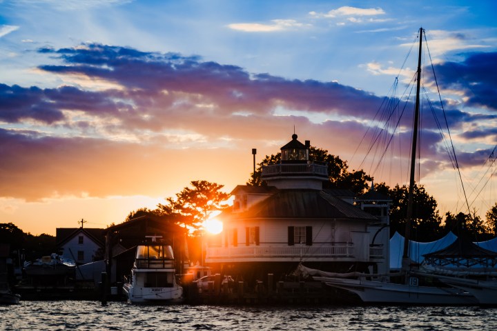 Sunset behind a lighthouse by the water with boats and trees silhouetted.