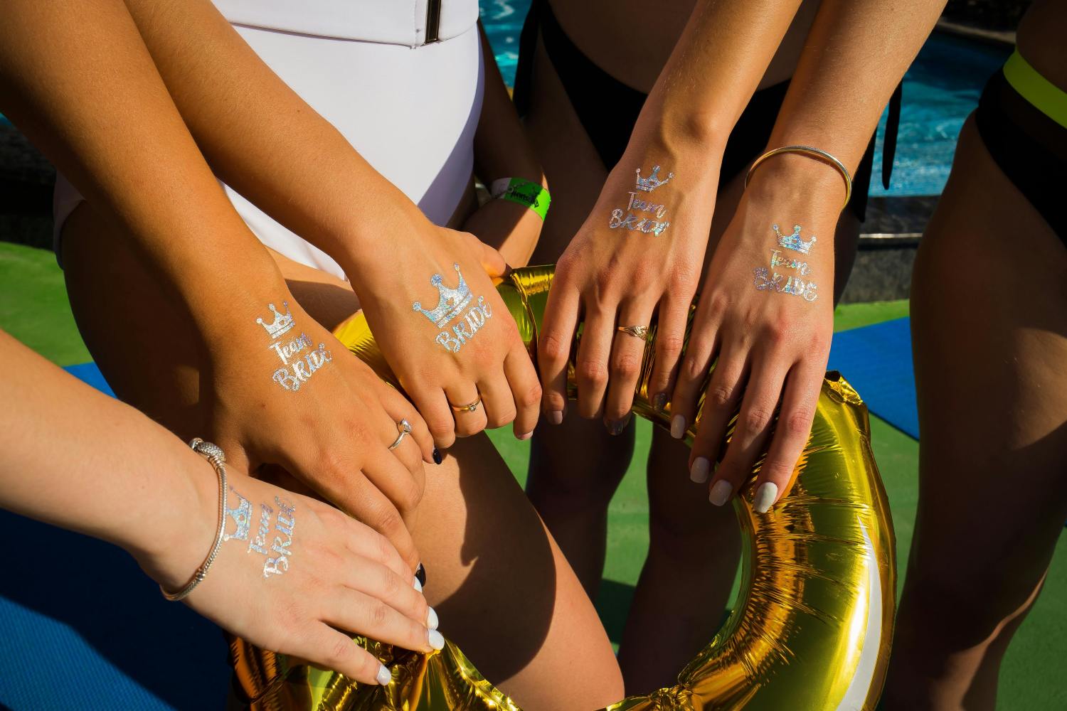 Five hands with 'Team Bride' tattoos holding a gold balloon by a pool.
