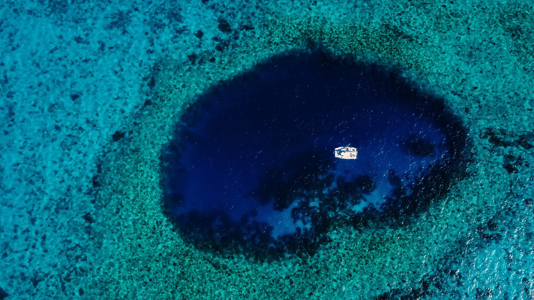Aerial view of a boat floating in a dark blue circular ocean spot surrounded by lighter turquoise water.