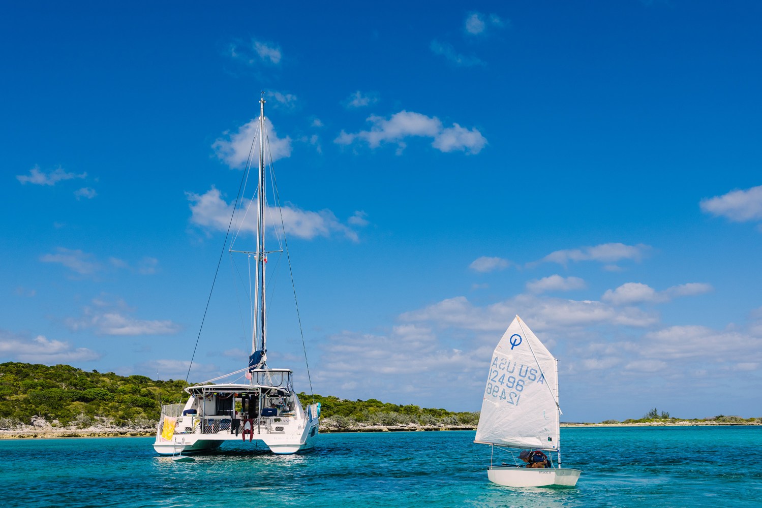 Sailboat and dinghy on turquoise water near lush green island under blue sky.