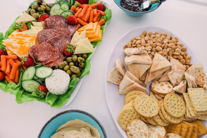 Two platters with meats, cheeses, veggies, crackers, nuts, and dip on a white table.