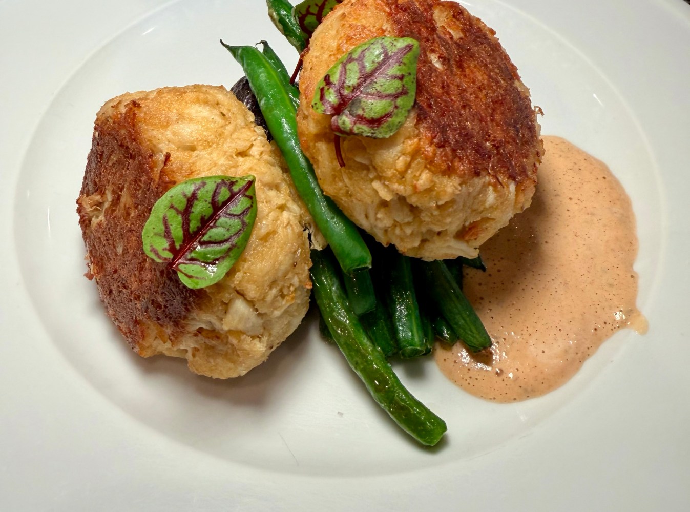 Two crab cakes on green beans with sauce and garnish on a white plate.