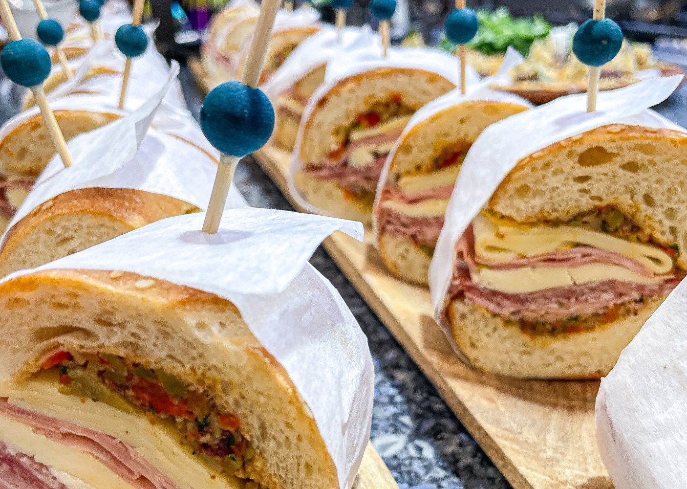 Sandwiches with deli meats and cheese on a board, skewered with toothpicks and wrapped in paper.