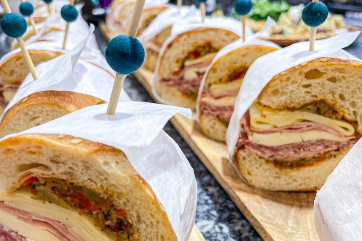Sandwiches with deli meats and cheese on a board, skewered with toothpicks and wrapped in paper.