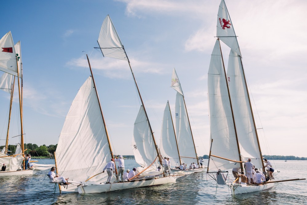Group of sailboats with white sails on a sunny day, people onboard navigating.