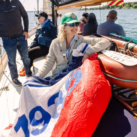 Smiling woman on a sailboat holding a colorful sail with '1929' on it.