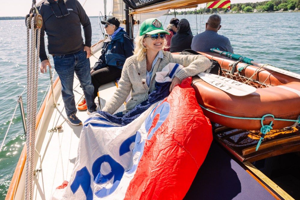 Smiling woman on a sailboat holding a colorful sail with '1929' on it.