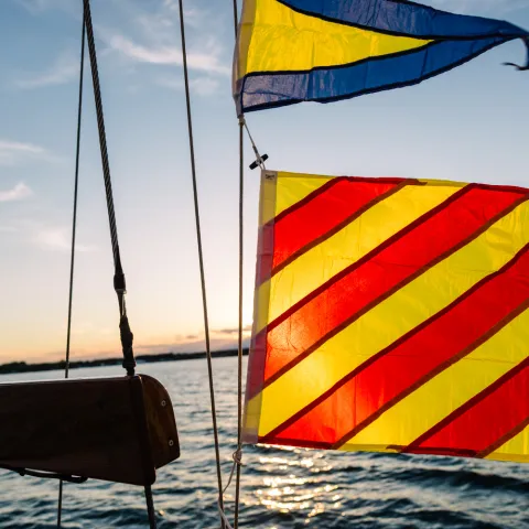 Sailboat flags with red and yellow stripes against a sunset on the water.