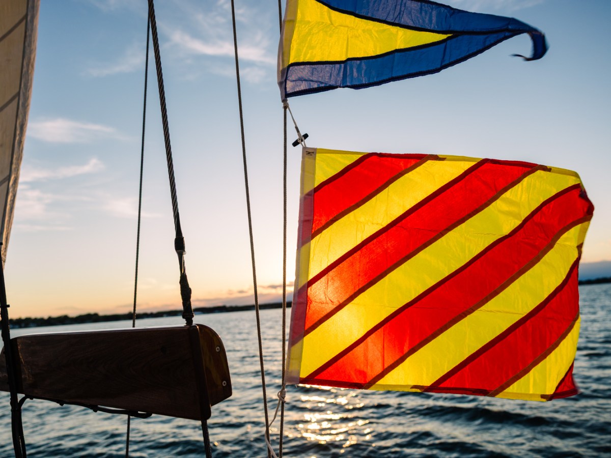 Sailboat flags with red and yellow stripes against a sunset on the water.