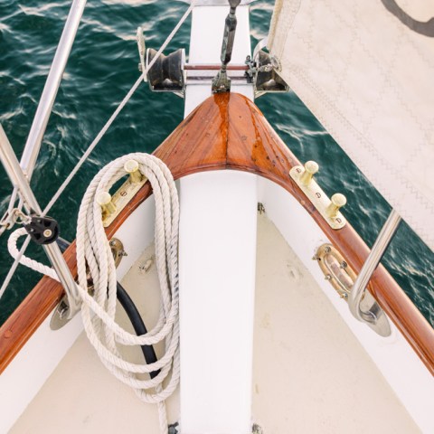 Sailboat bow with ropes and a sail visible on calm water.