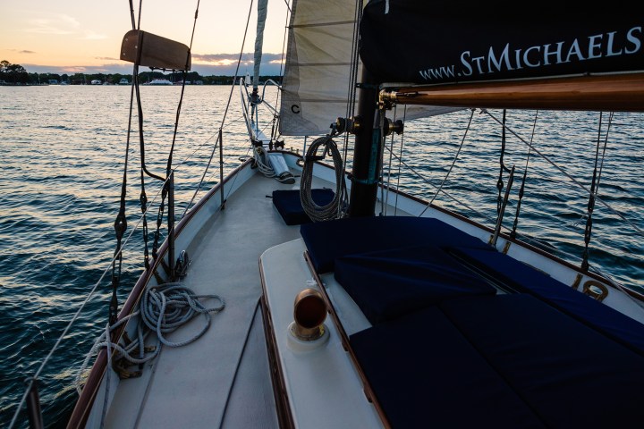 Sailboat deck on calm water at sunset, with sails and ropes visible.