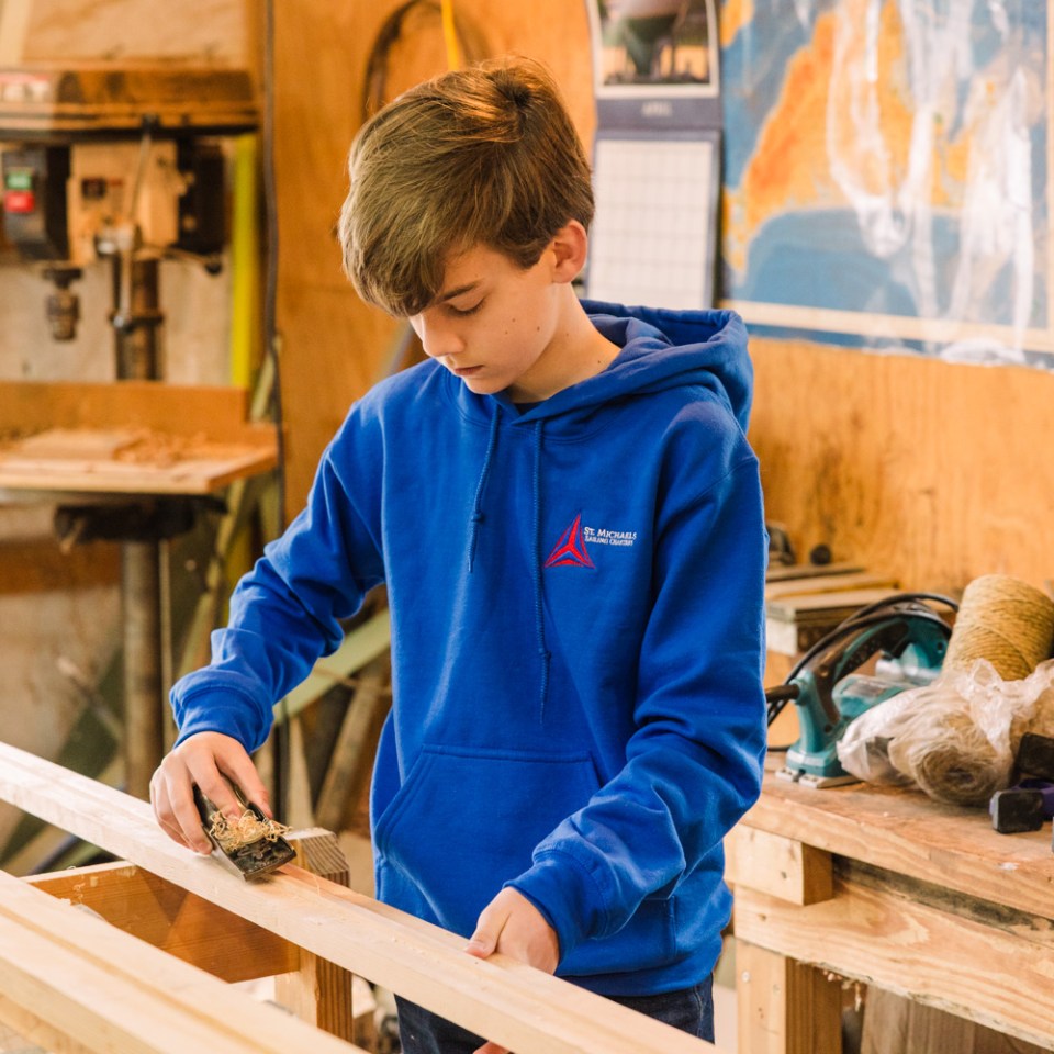 Boy in blue hoodie sanding wood in a workshop.