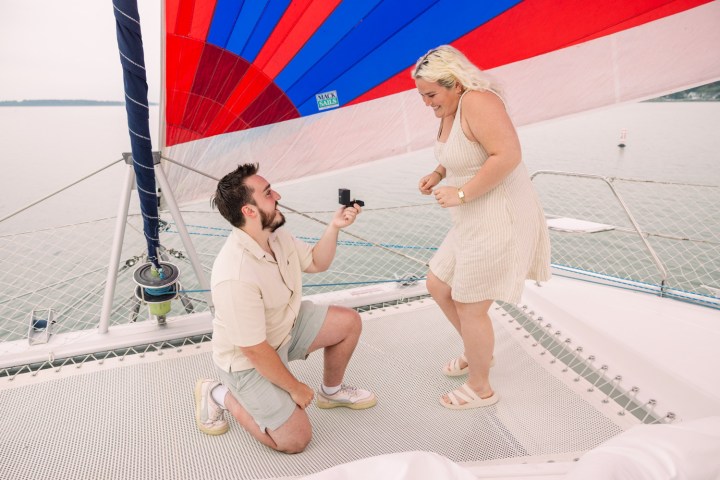 Person kneeling and proposing on a sailboat under a colorful sail.