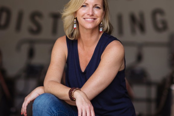 Woman smiling, sitting on a stool in a casual pose with blurred distillery background.