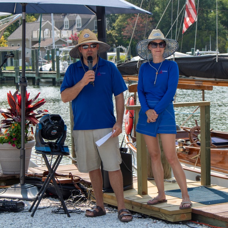 Two people in blue shirts and hats speaking by a marina.