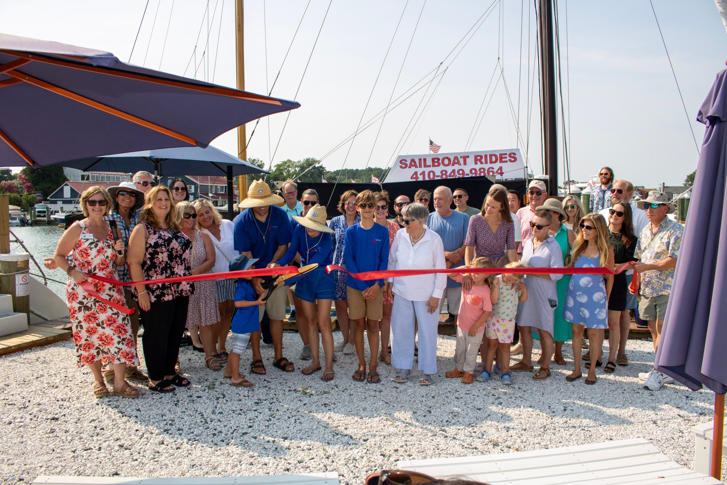 Group at a dockside event cutting a red ribbon for sailboat rides.