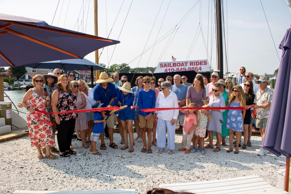Group at a dockside event cutting a red ribbon for sailboat rides.