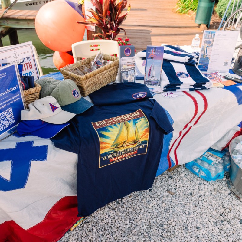 Table display with shirts, hats, flyers, and a fan on a flag tablecloth near a dock.