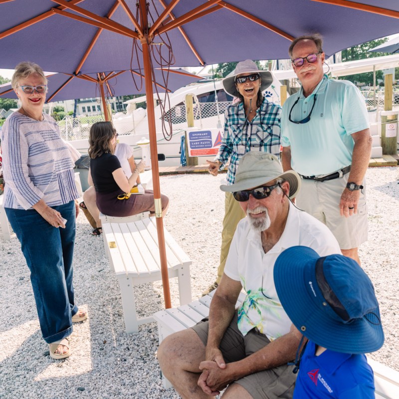 A group of people under umbrellas at an outdoor event with gravel ground and a musician in the background.