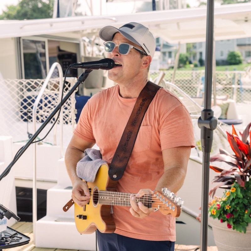 Man in sunglasses sings and plays guitar near a boat.
