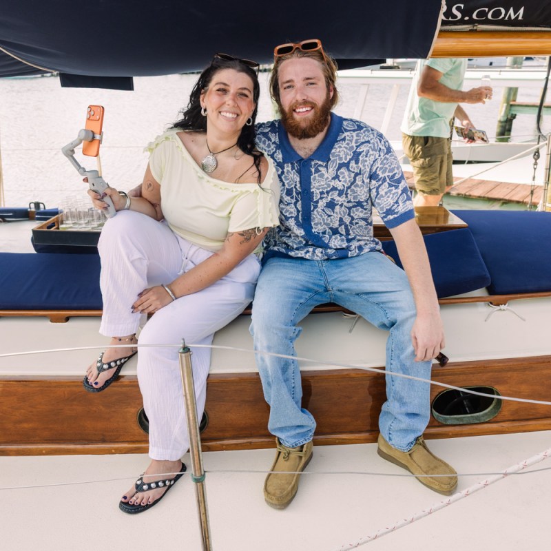 Two people smiling on a boat deck, one holding a bottle, near a marina.