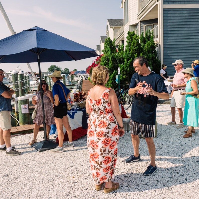 People socializing outdoors near a marina, with a boat and umbrellas in the background.