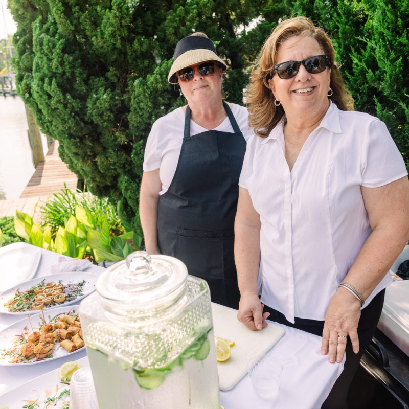 Two women in sunglasses by a table with food and drinks near a marina.