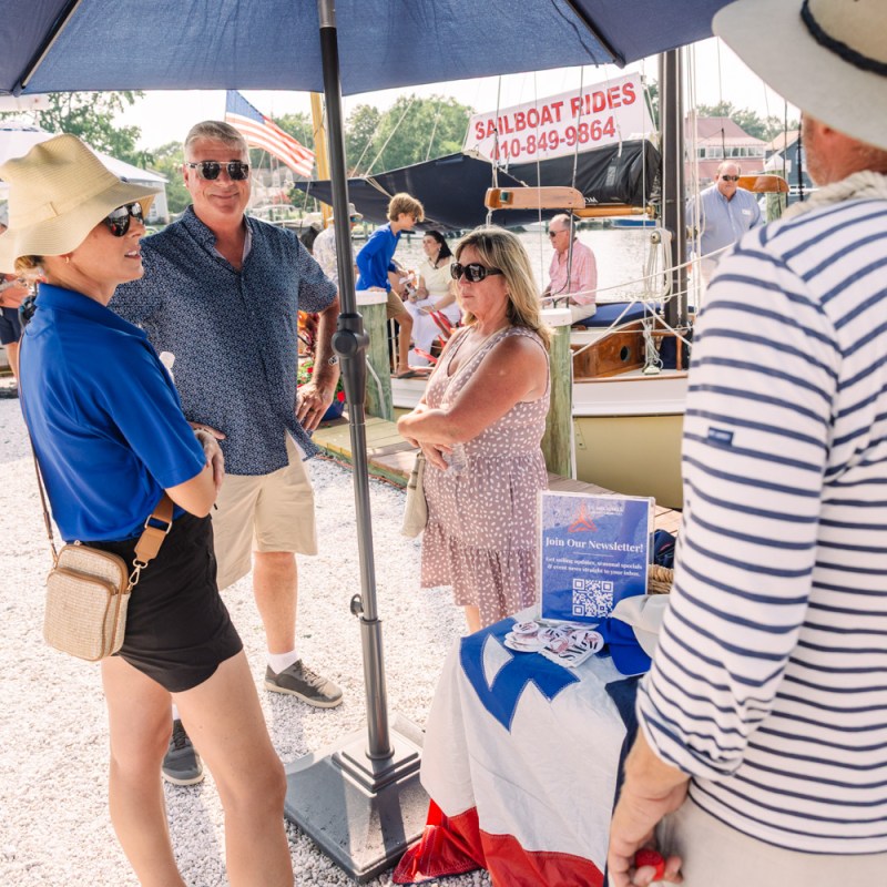People chatting at a marina booth under an umbrella with sailboat ride info.