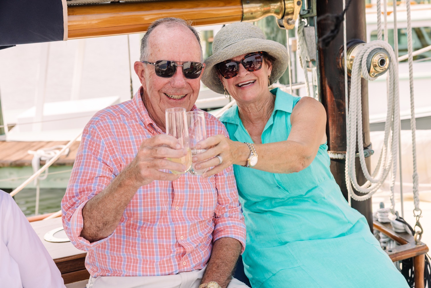 Older couple smiling, toasting drinks on a boat, wearing sunglasses and summer clothing.