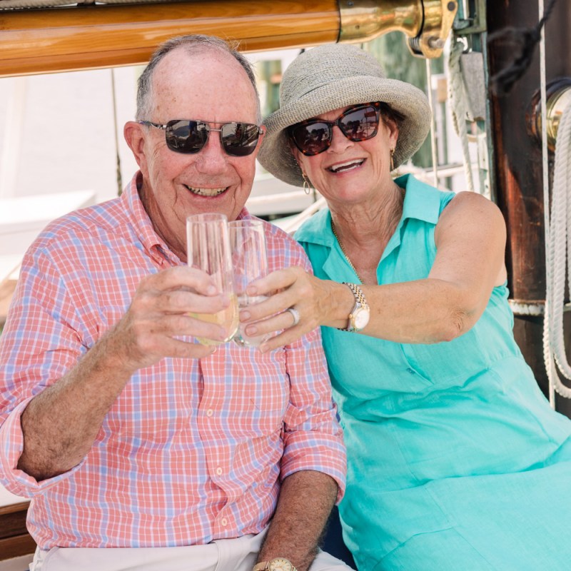 Older couple smiling, toasting drinks on a boat, wearing sunglasses and summer clothing.