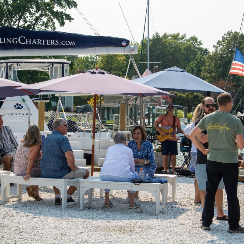 People sitting and standing outdoors near boats with umbrellas and a musician.