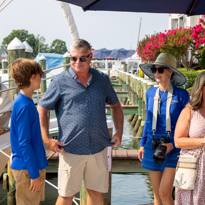 Four people stand on a sunny marina dock near moored boats.