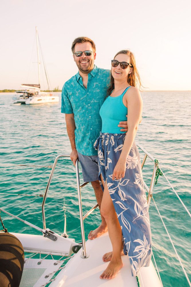 Couple smiles on a sailboat at sunset, wearing sunglasses and casual summer clothes.